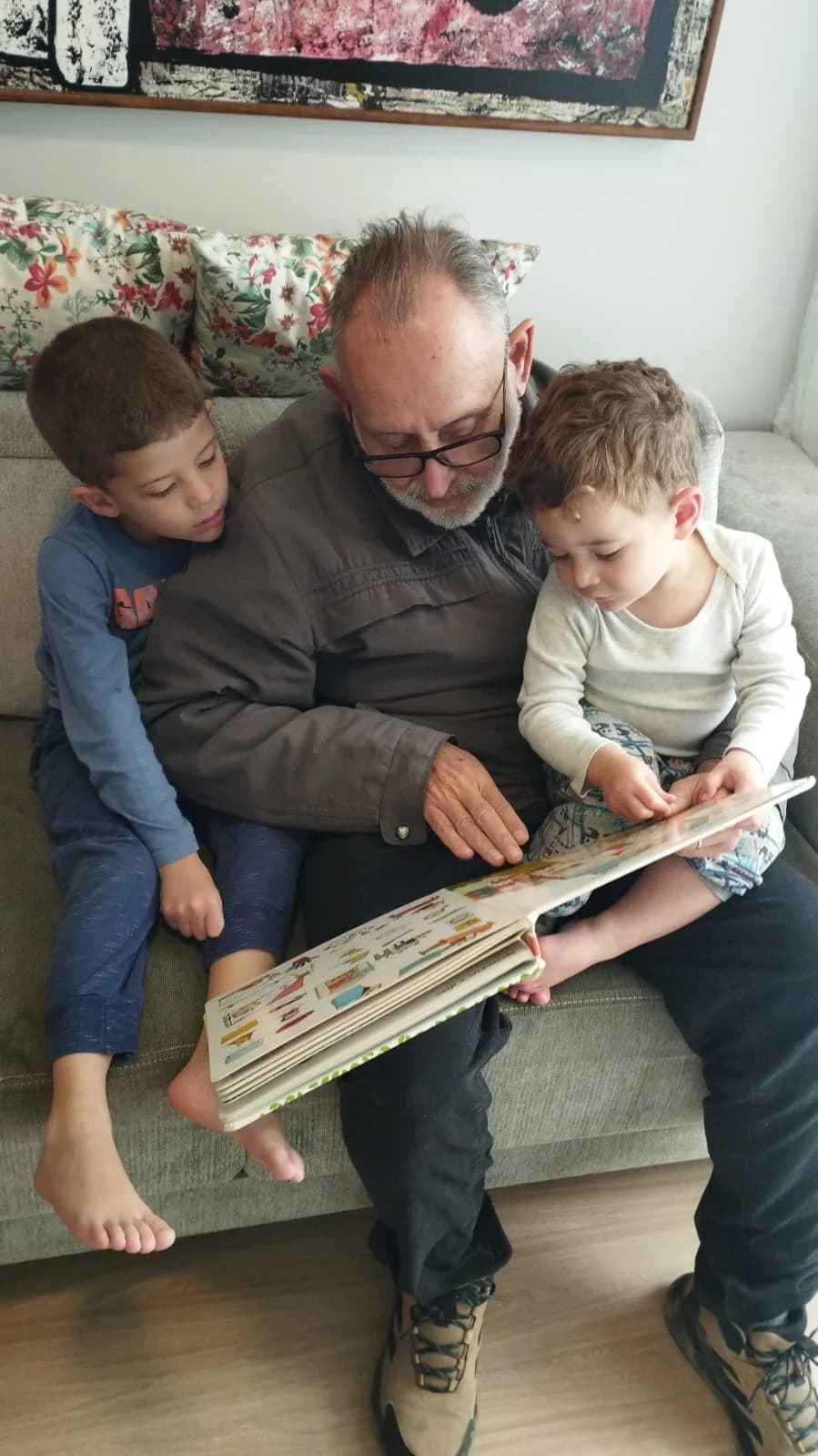 Silvio Roberto de Oliveira lendo um livro no sofá com seus dois netos.